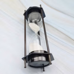 The hexagonal brass sand timer tilted to show the white sand flowing through the hourglass.