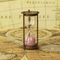 A small antique brass 1-minute hourglass with light pink sand, standing on a vintage world map.