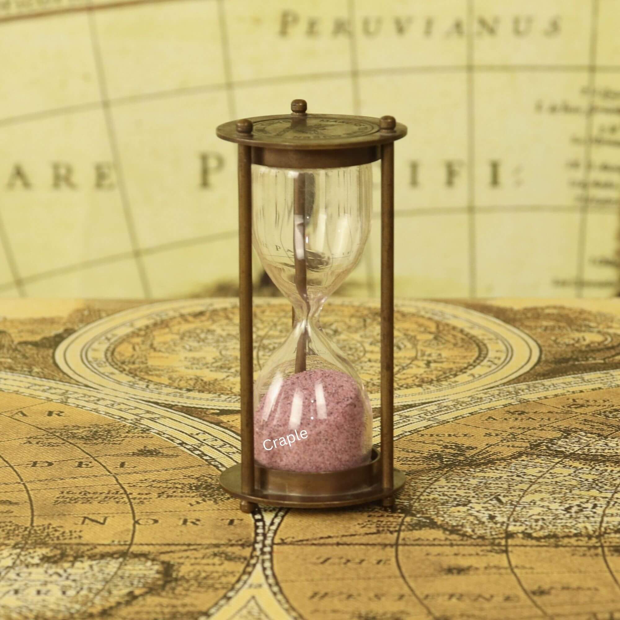Makers To The Queen Brass Antique SandTimer-1 A small antique brass 1-minute hourglass with light pink sand, standing on a vintage world map.