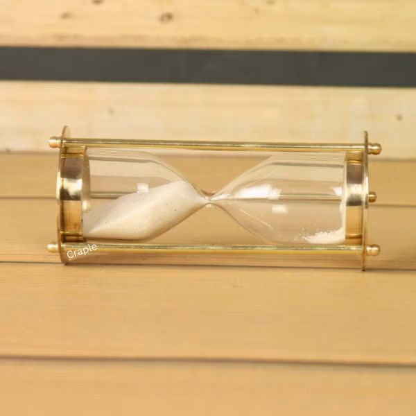 The miniature brass hourglass resting horizontally on a wooden surface, with white sand settled in the glass bulb.