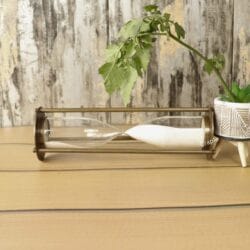 The slim antique brass hourglass resting horizontally on a wooden table, with white sand settled inside.