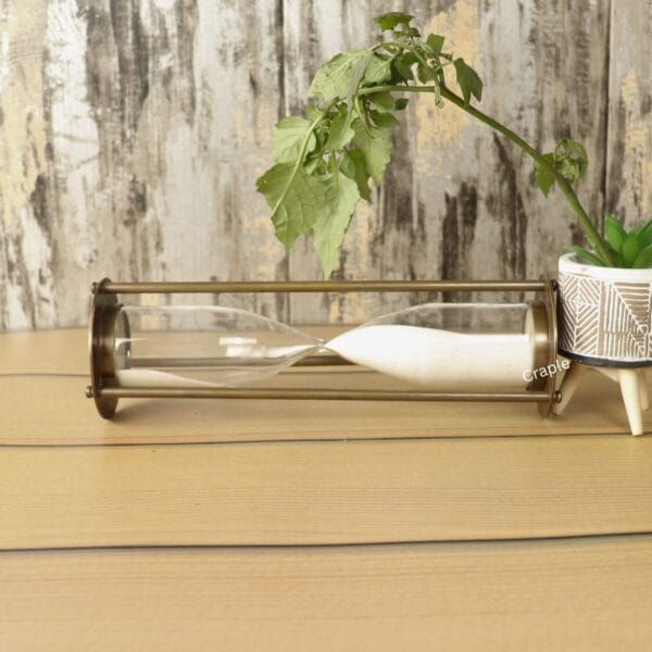 The slim antique brass hourglass resting horizontally on a wooden table, with white sand settled inside.