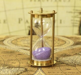 A petite 1-minute hourglass with a shiny brass frame and purple sand, displayed on a vintage map.