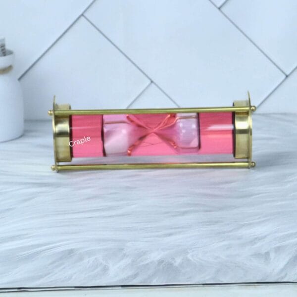 The decorative pink liquid hourglass lying horizontally, showcasing the floating sand timer within its brass frame.