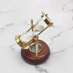 Rotating brass hourglass with white sand and a built-in compass, mounted on a round, polished wooden base.