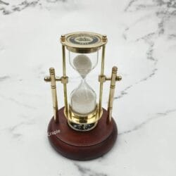 Upright brass sand timer and compass with a polished wood base against a marble background.