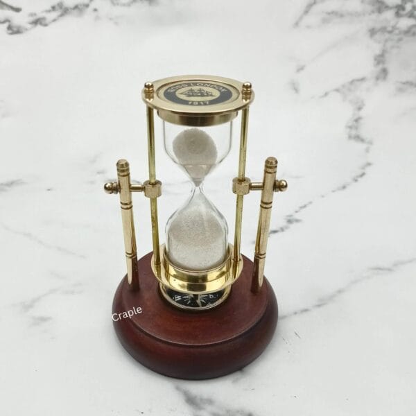 Upright brass sand timer and compass with a polished wood base against a marble background.