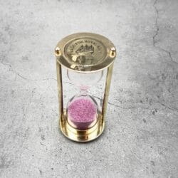 Top-down view of a Royal Navy brass sand timer with pink sand, showing the engraved top plate.