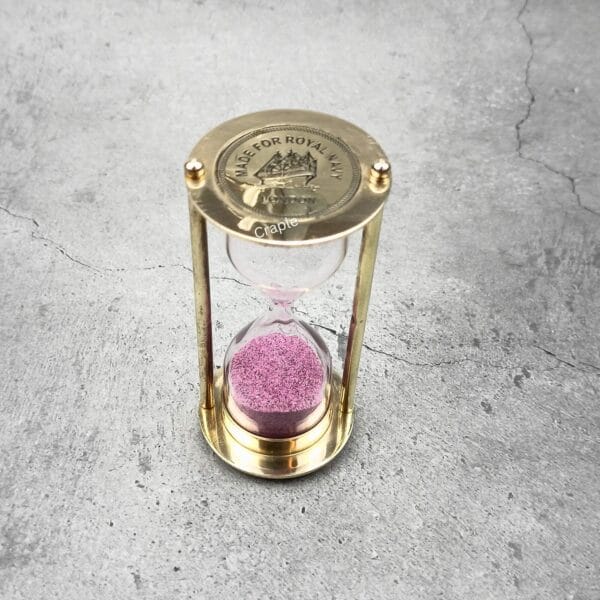 Top-down view of a Royal Navy brass sand timer with pink sand, showing the engraved top plate.