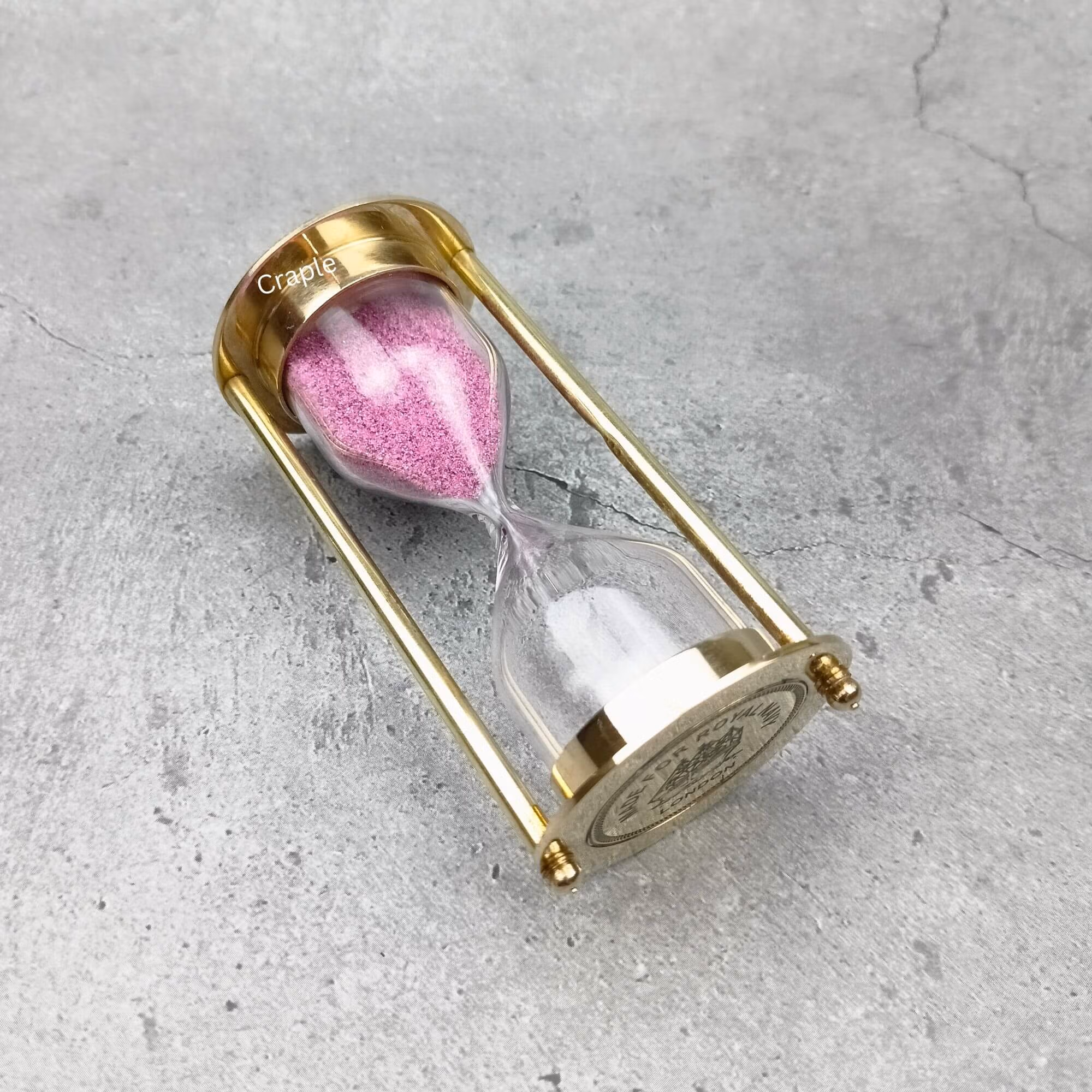 A 3-minute Royal Navy hourglass with a polished brass frame and pink sand, resting on its side to showcase its design.