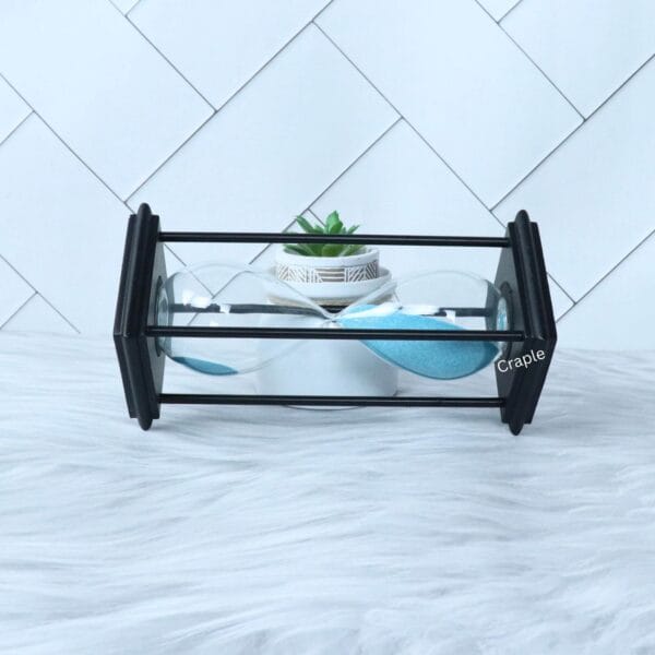 The modern blue sand hourglass displayed as a decorative piece on a white surface next to a plant.