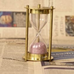 Elegant brass 3-minute hourglass sand timer with shimmering pink sand, standing upright on a newspaper.