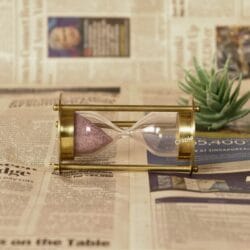 The brass and pink sand hourglass lying on its side on a vintage-style newspaper background.