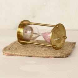 Decorative brass and pink sand timer displayed at an angle on a small rustic burlap mat.