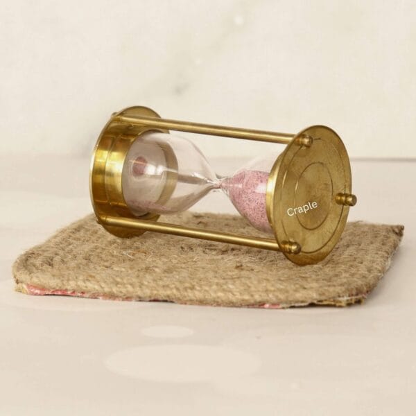 Decorative brass and pink sand timer displayed at an angle on a small rustic burlap mat.
