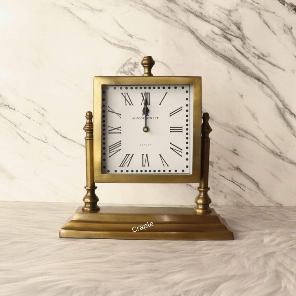 A square tilting table clock with an antique brass finish, featuring a white dial with Roman numerals, set on a sturdy decorative stand.